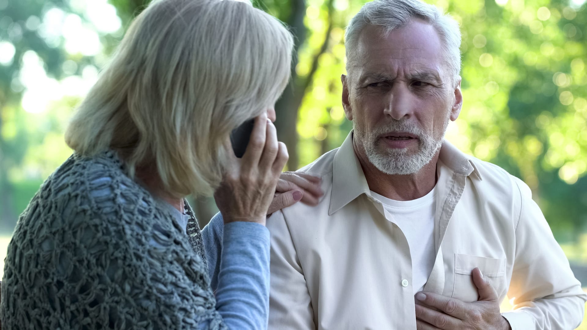 Mann mit Brustschmerzen neben telefonierender Frau Mann mit Brustschmerzen neben telefonierender Frau